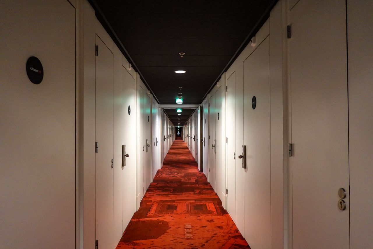 our-story Long, symmetrical hotel hallway with red carpet and closed white doors under warm lighting.